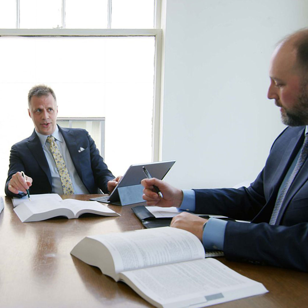 Two man wearing a suit holding a pen with books on top of the table